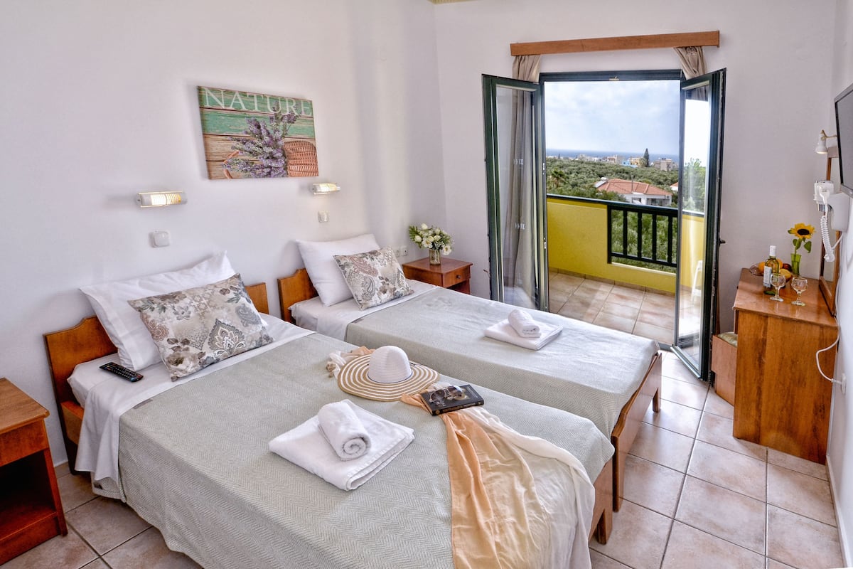A hotel room with two beds, a balcony with a view, a painting on the wall, and a small table with a vase and a phone.