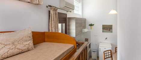 A bedroom with a wooden bed, a small kitchenette, a refrigerator, and a dining area with a chair.