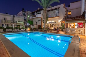 A swimming pool with lounge chairs and umbrellas, surrounded by buildings with balconies and outdoor seating areas. - Galini Apartments and Studios (Hersonissos)