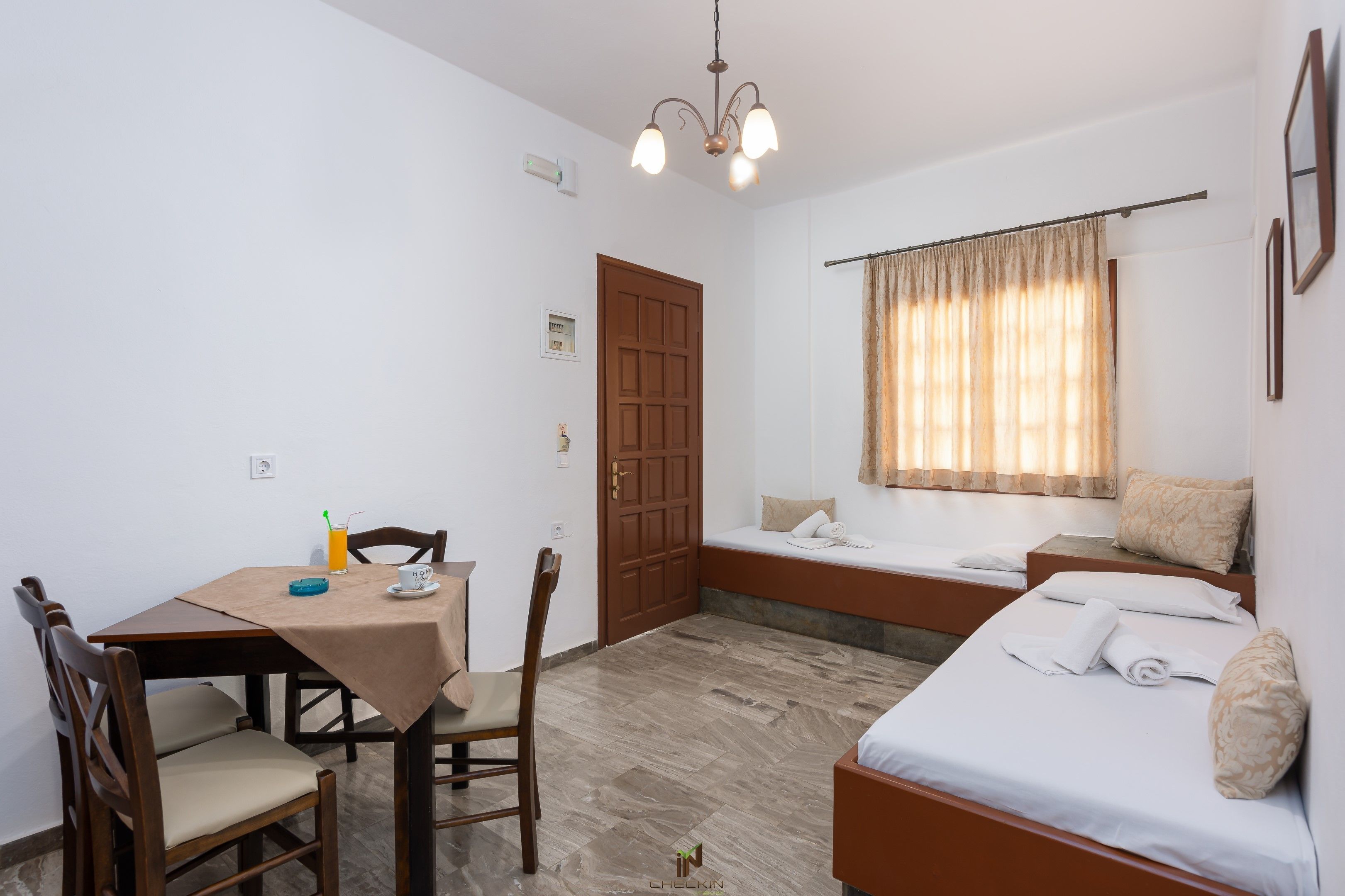 A room with a bed, a small table with a chair, a bench, and a window with curtains.