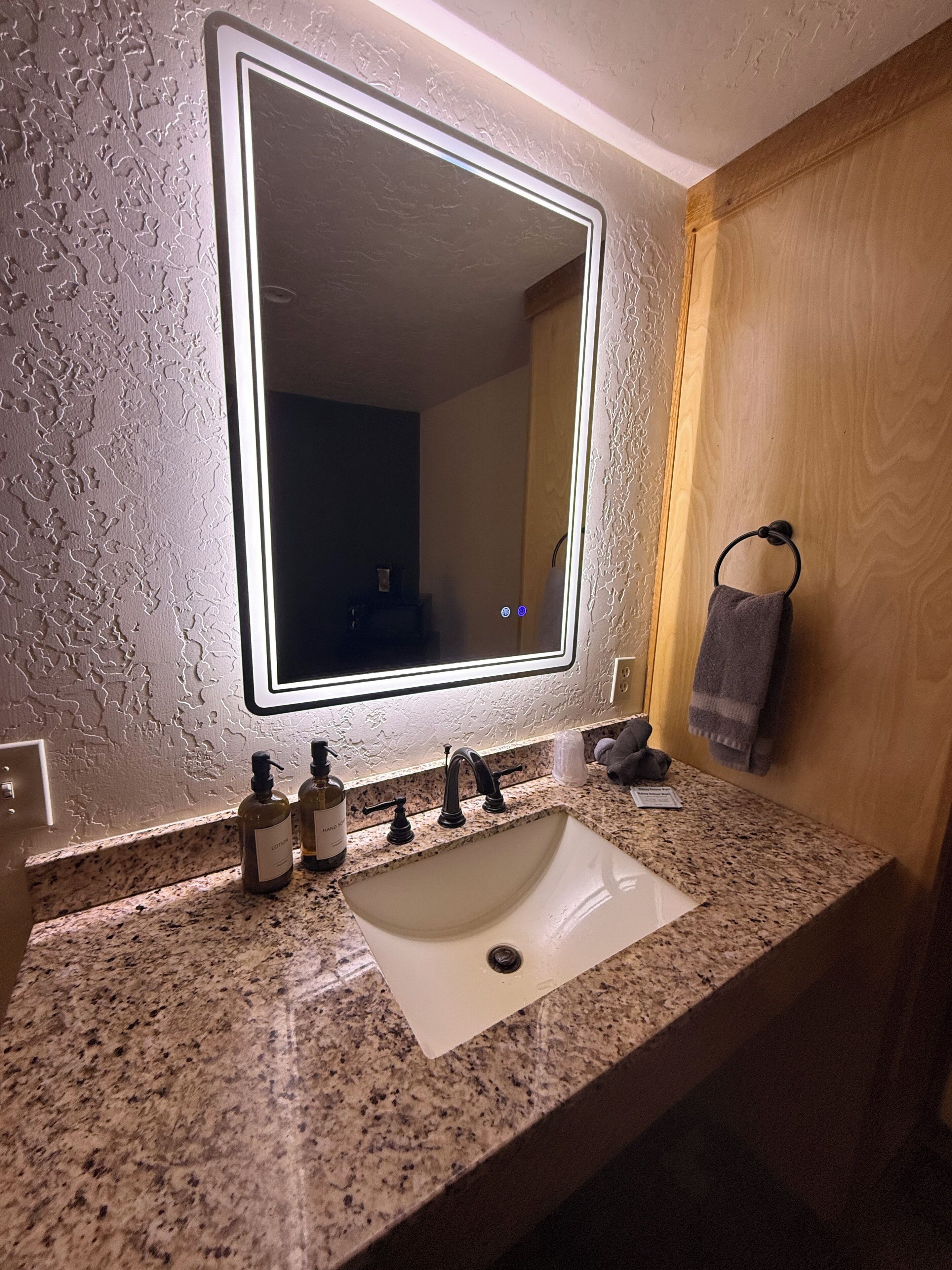 A bathroom with a granite countertop, a white sink, a mirror with a light, and a towel rack.