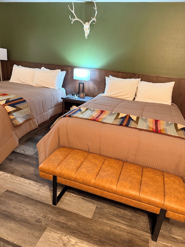 A hotel room with two beds, a bench, a nightstand with a lamp, and a wall-mounted deer head.