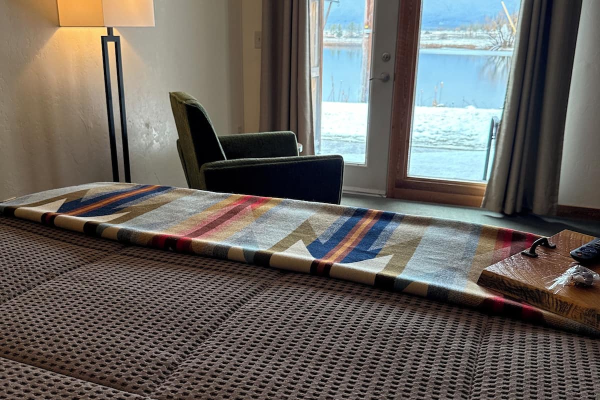 A hotel room with a large bed, a chair, a lamp, and a view of a lake through a sliding door.