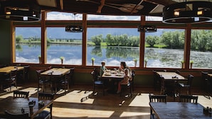 A restaurant with large windows overlooking a lake, wooden tables and chairs, and hanging lights.