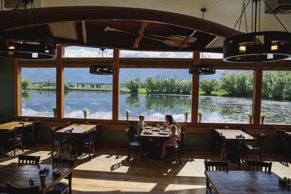 Un restaurant avec de grandes fenêtres donnant sur un lac, des tables et des chaises en bois, et des luminaires suspendus.