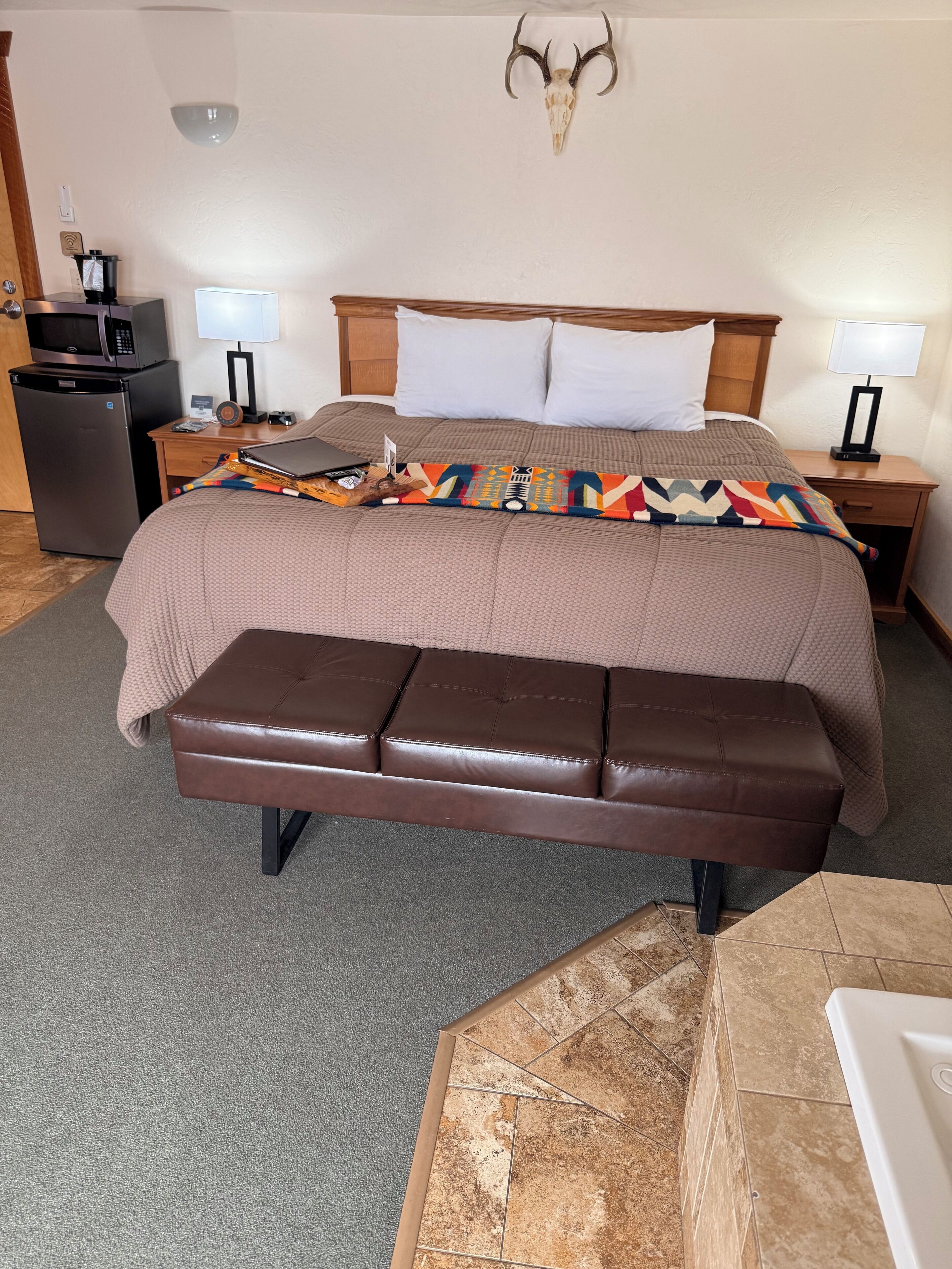 A hotel room with a bed, a wooden headboard, a mounted deer skull, a desk with a computer, a refrigerator, and a bench.
