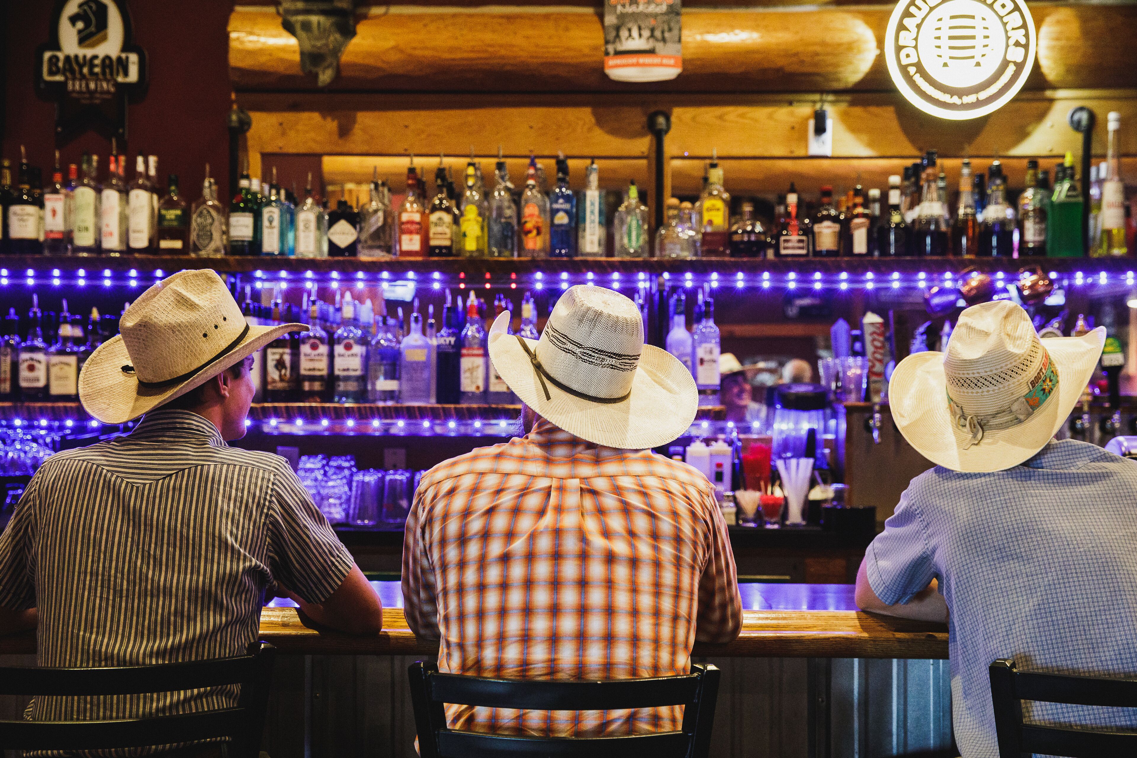 Three men seated at a bar with a variety of liquor bottles in the background.