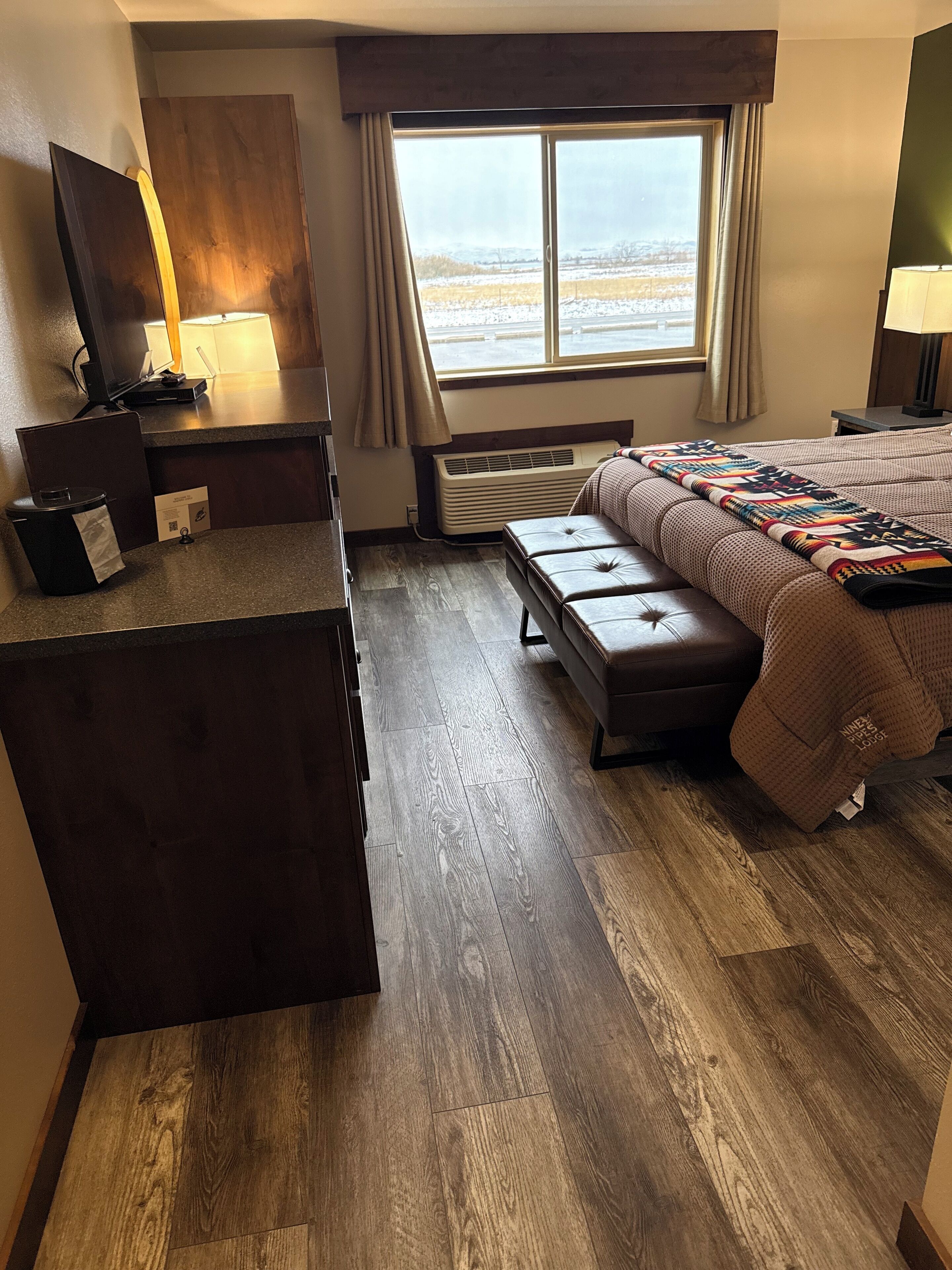 A hotel room with a large bed, a desk with a computer, a window with a view of a snowy landscape, and a heater under the window.