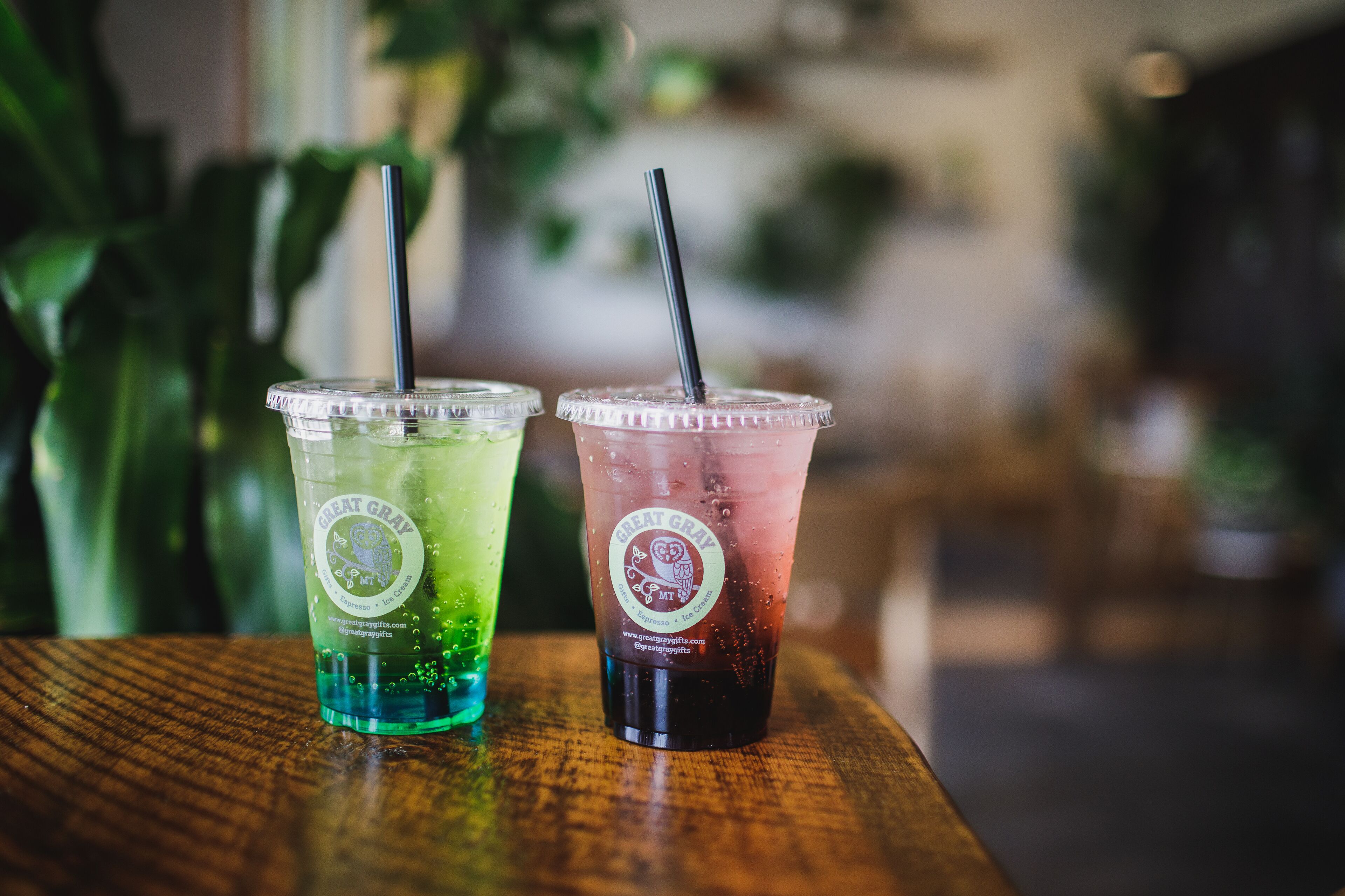 Two iced drinks in clear plastic cups with black straws on a wooden table.