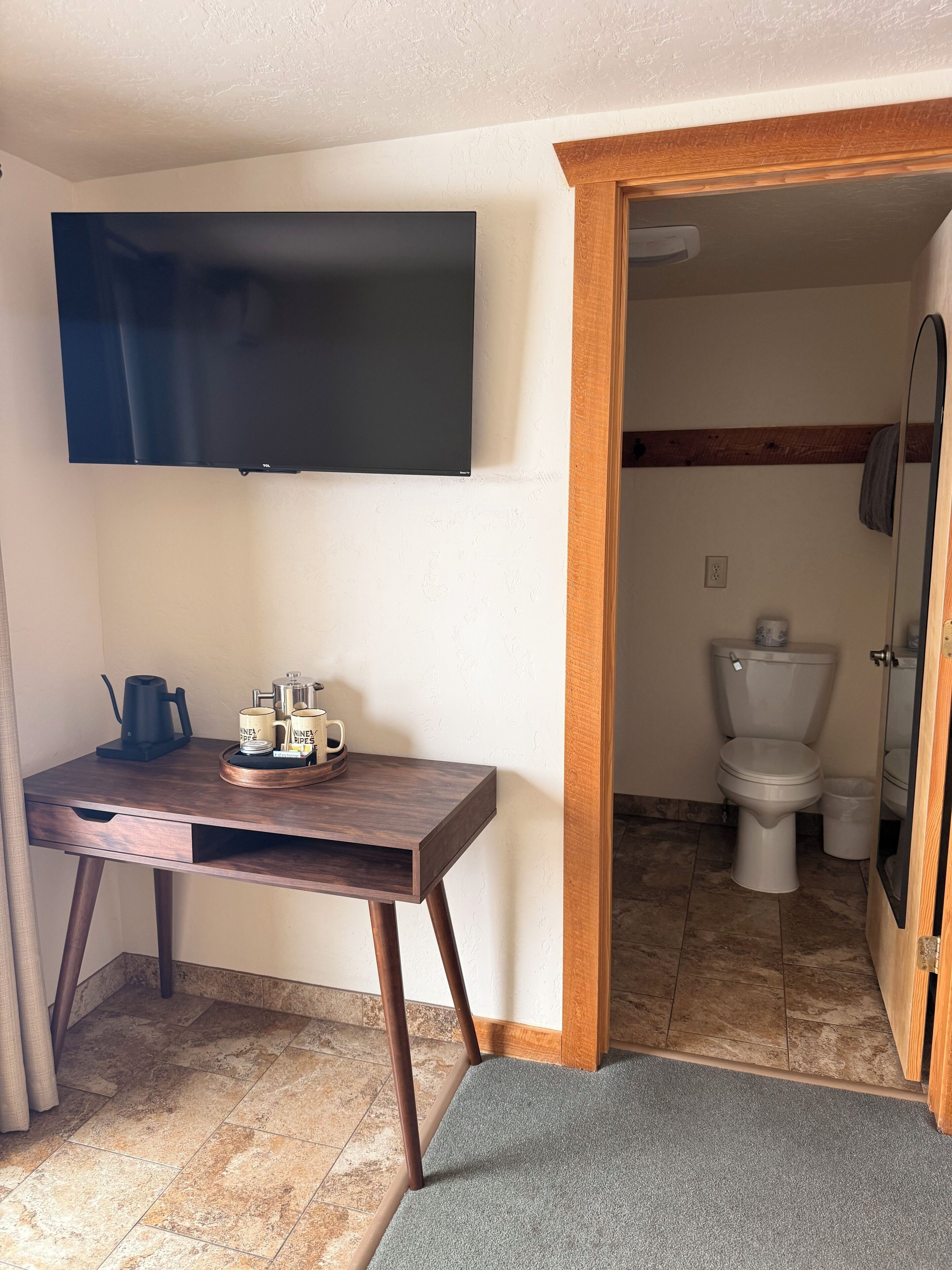 A small room with a wooden desk, a flat-screen TV mounted on the wall, and a bathroom visible through an open door.