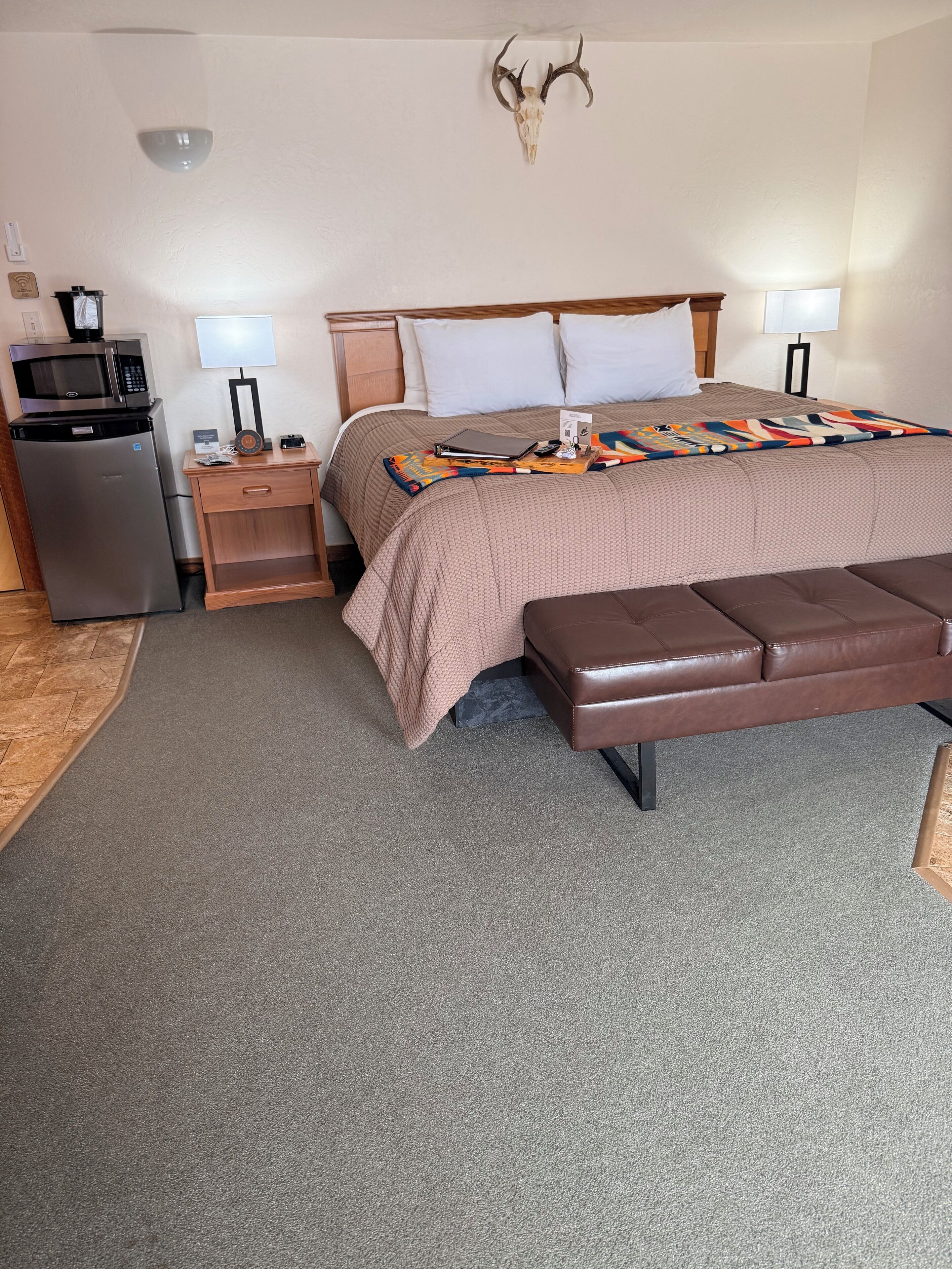 A hotel room with a bed, a desk with a microwave, a refrigerator, a wall-mounted deer skull, and a patterned blanket.