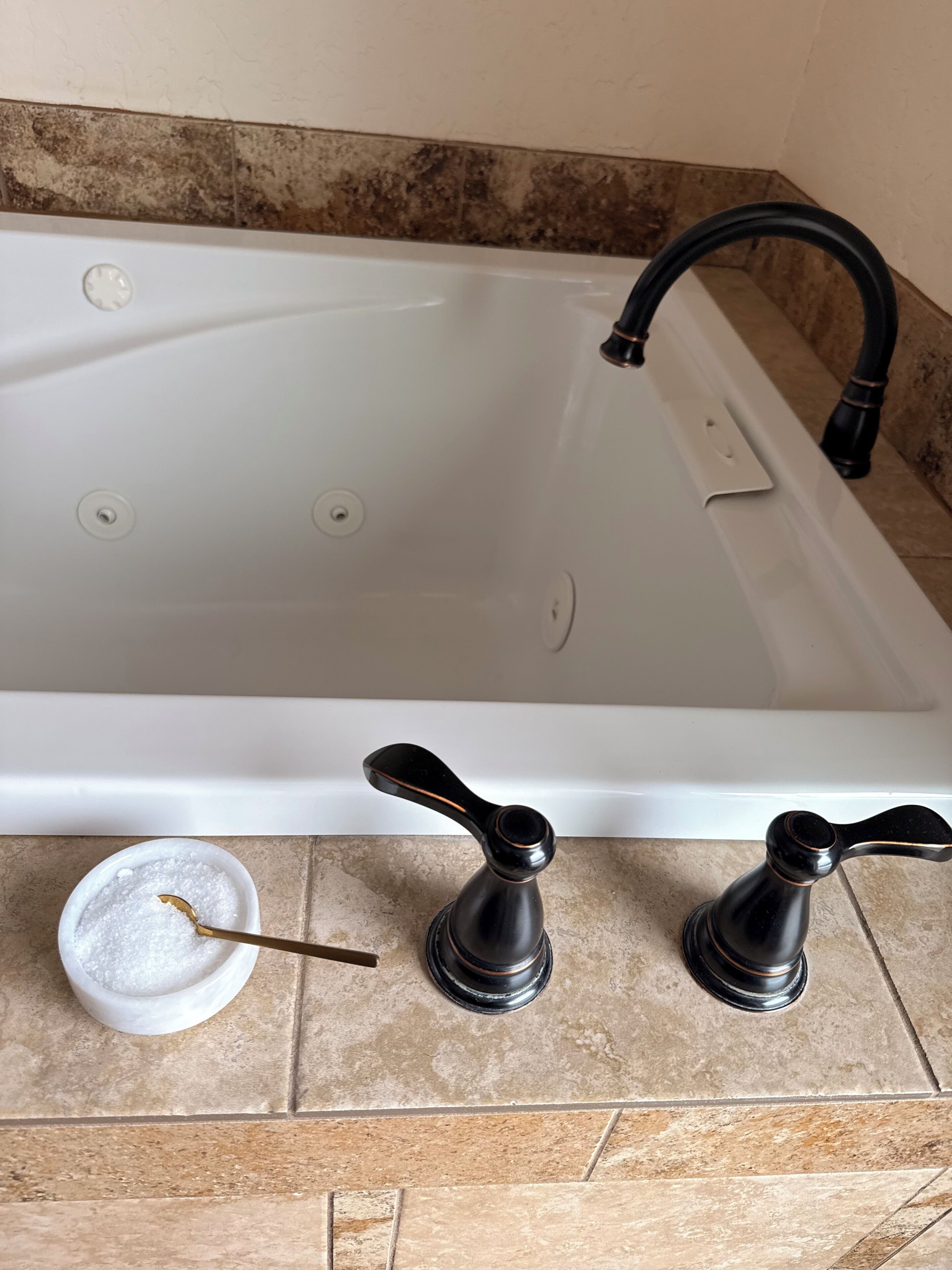 A bathtub with a black faucet and two black handles, a white basin with a gold spoon, and a beige tiled wall.