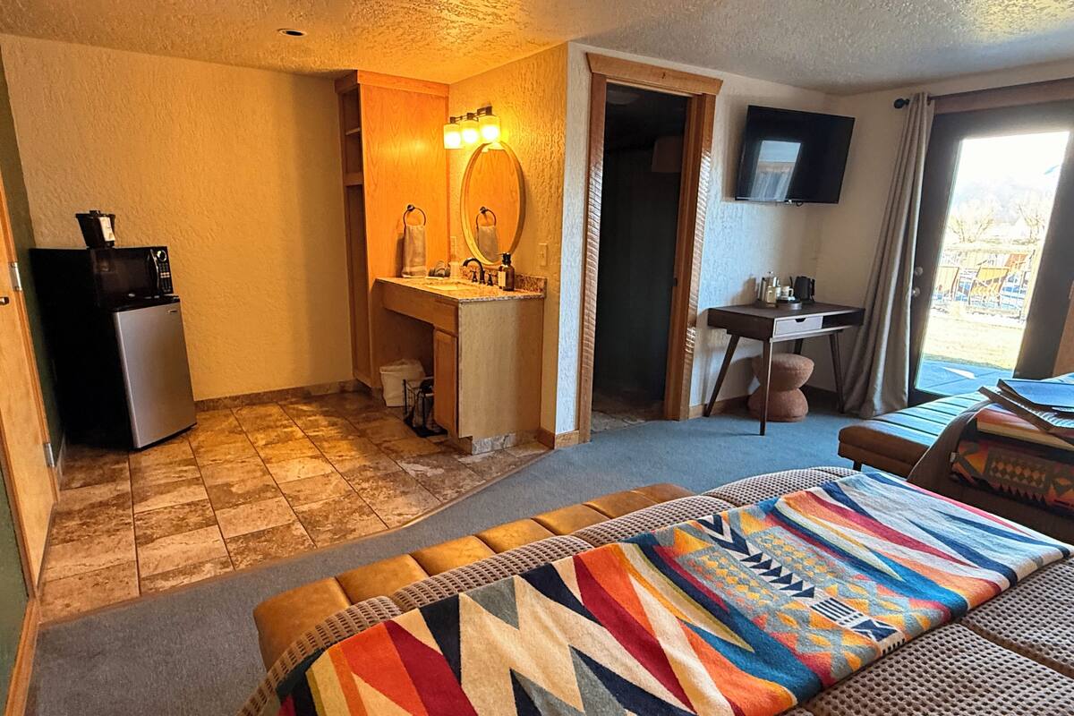A hotel room with a bed, a small desk, a bathroom with a sink and mirror, and a television on the wall.