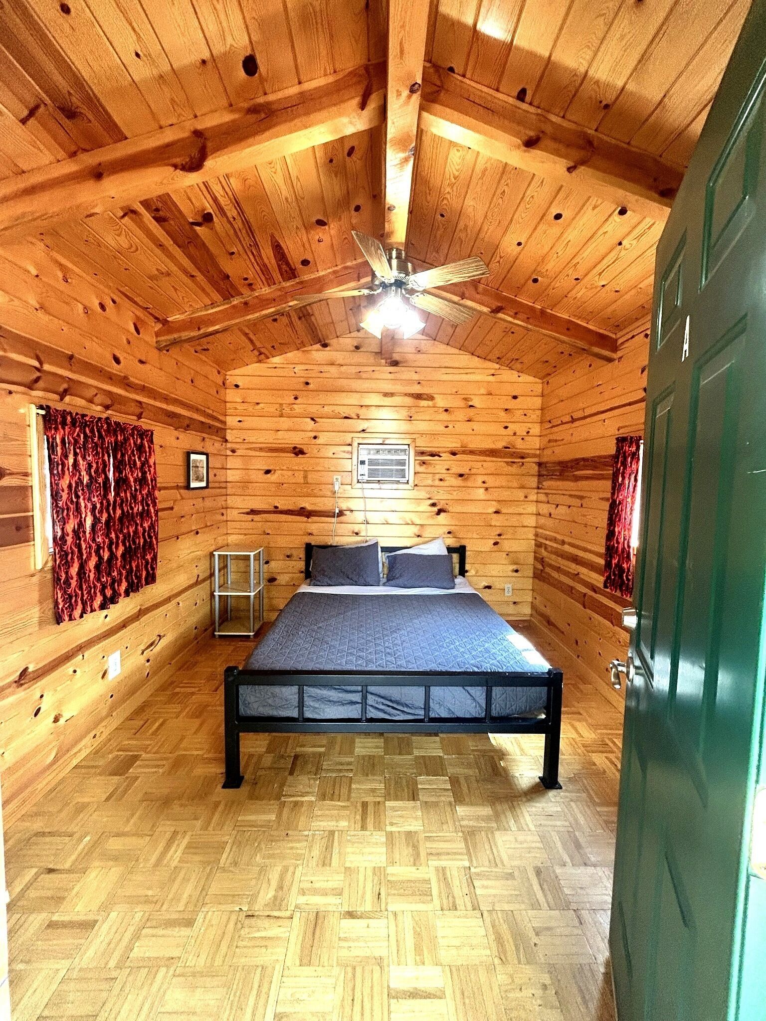A wooden cabin room with a bed, a ceiling fan, and a window with curtains.