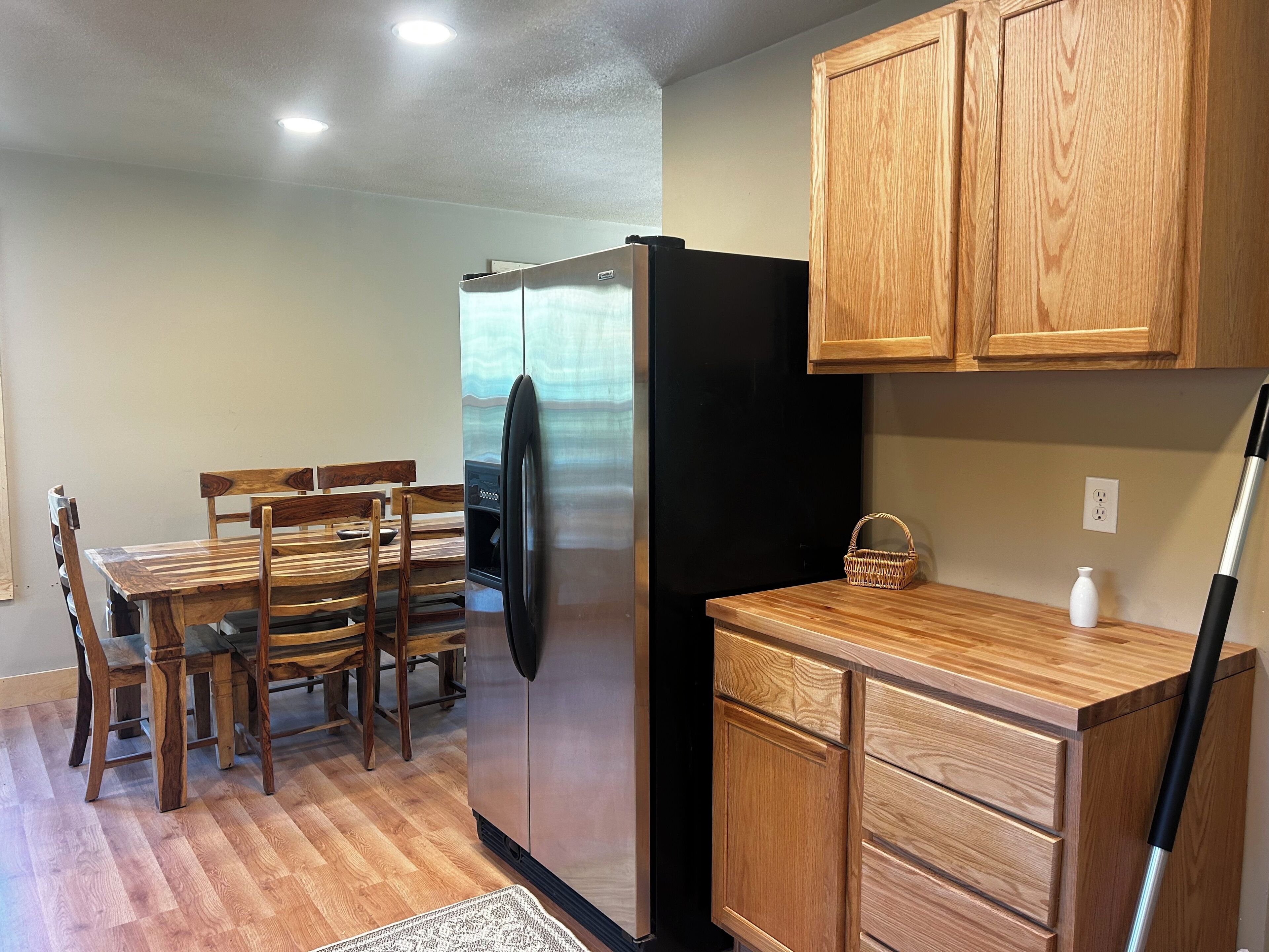 A compact kitchen with wooden cabinetry, a stainless steel refrigerator, a dining table with chairs, and a small countertop with a basket and a vase.