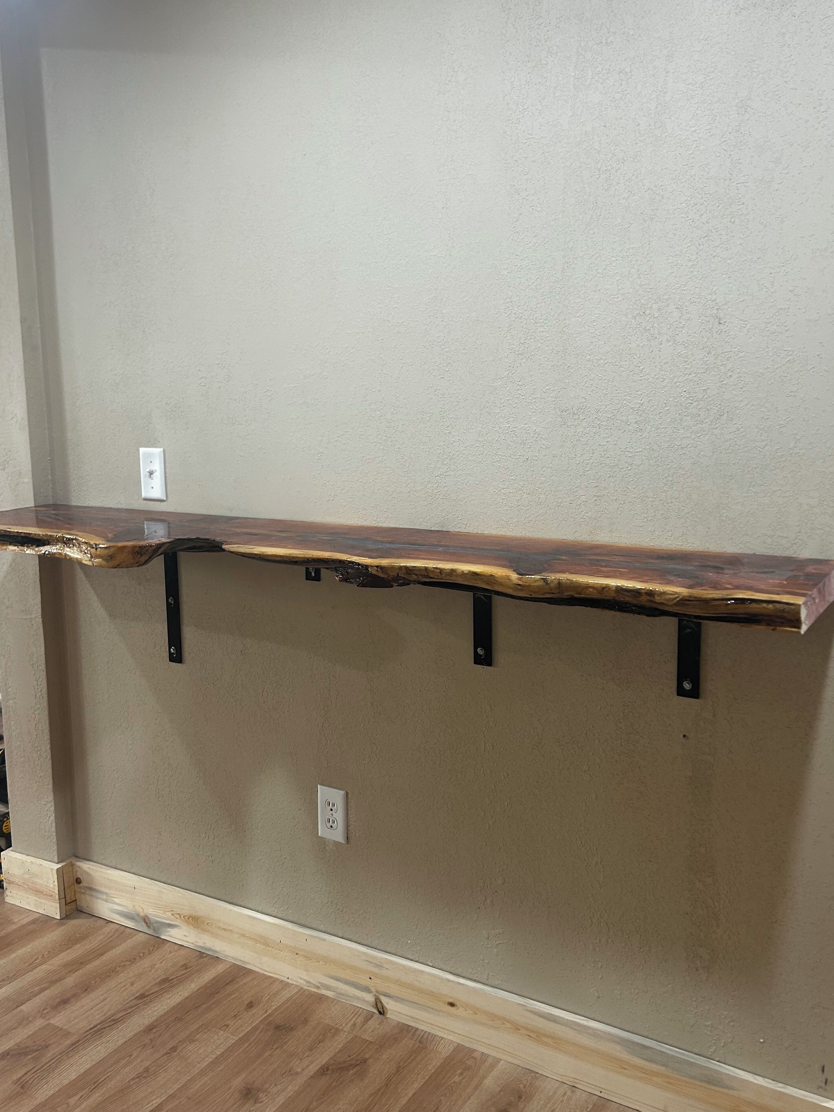 A wooden shelf with metal brackets against a textured wall, with a power outlet below.
