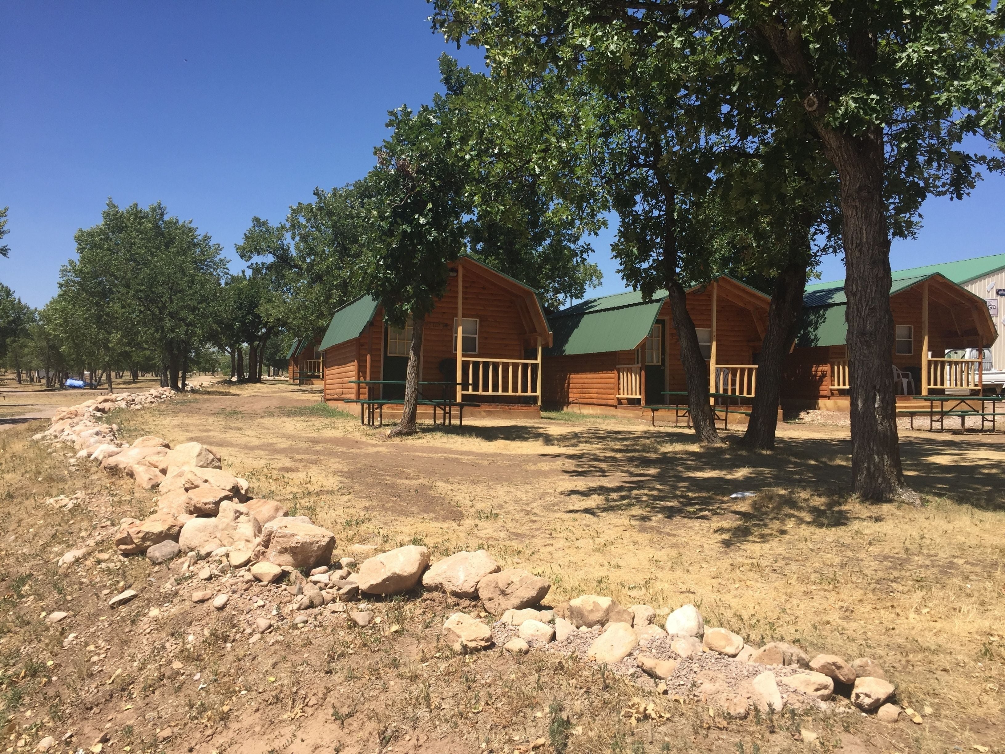 A row of wooden cabins with green roofs, surrounded by trees and a dirt path.