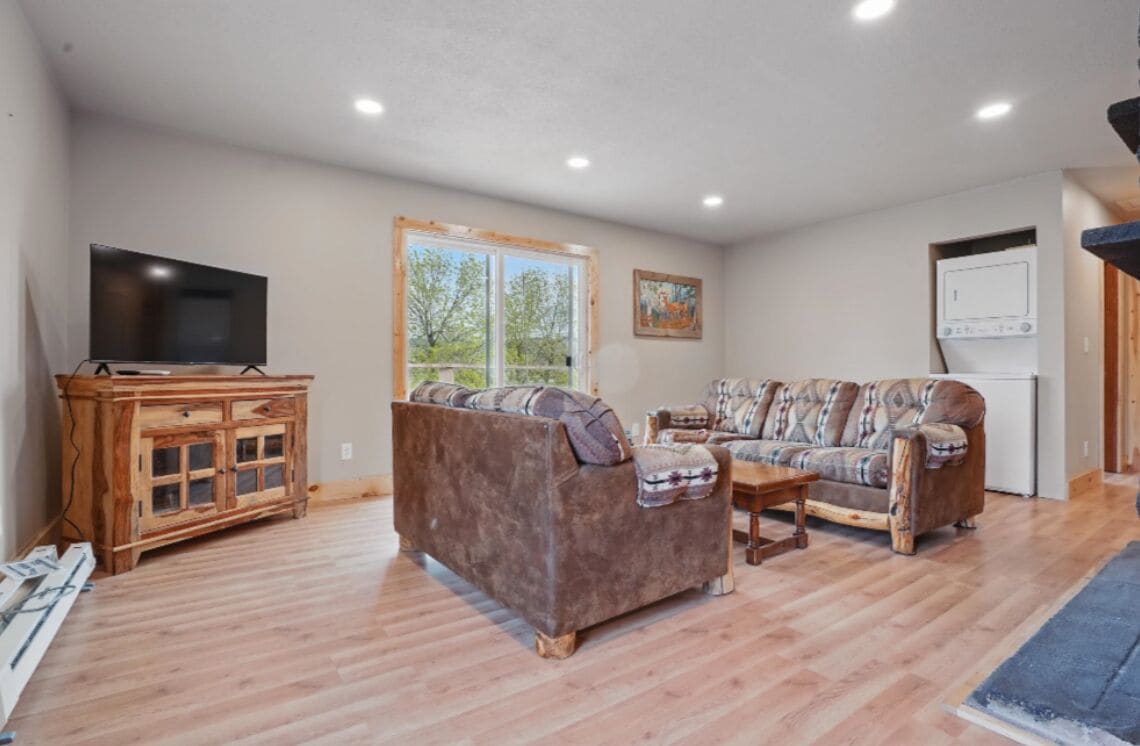 A living room with a large flat-screen TV, two sofas, a wooden cabinet, and a window with a view of trees.
