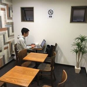 A person working on a laptop at a wooden table in a room with two windows, a no-smoking sign, and a potted plant.
