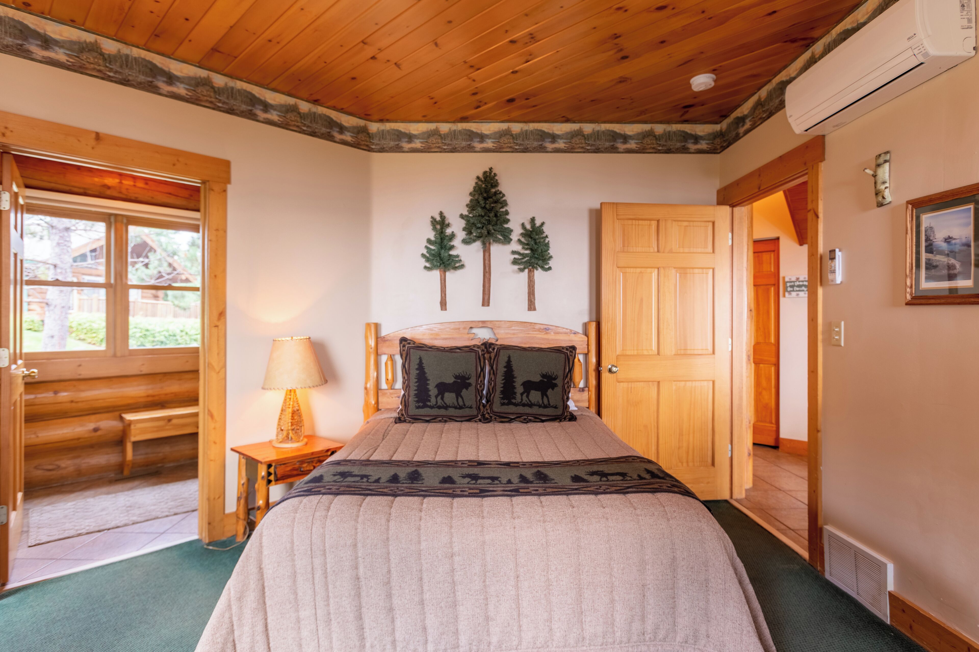 A bedroom with a wooden ceiling, a bed with a blanket, a nightstand with a lamp, a door, and a window.