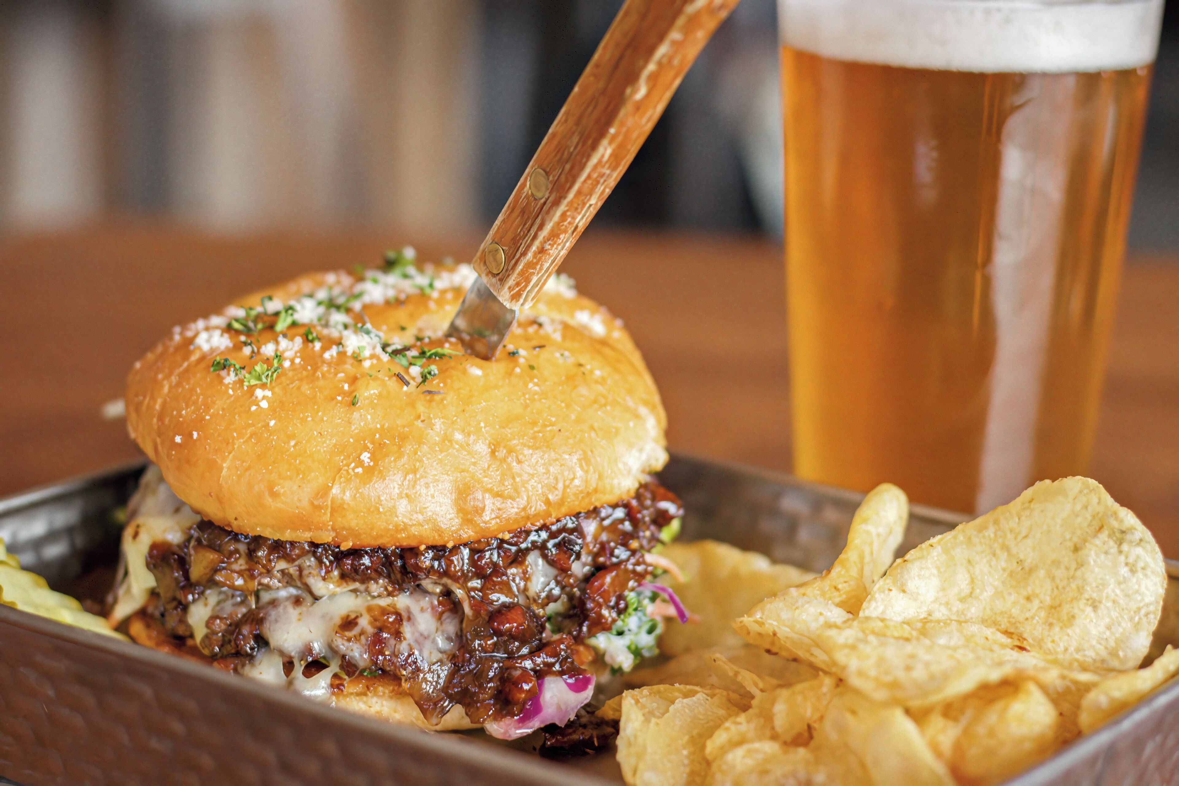 A burger with a fork, a glass of beer, and potato chips on a tray.