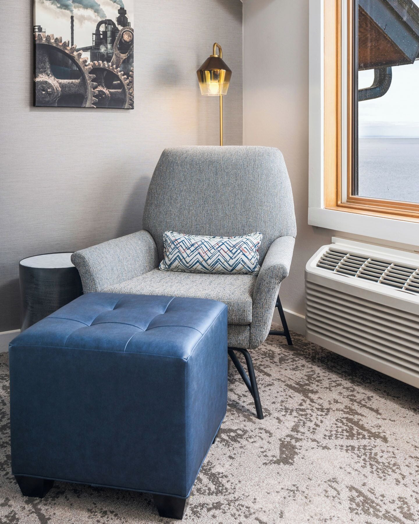 A modern living room with a grey armchair, a blue ottoman, a floor lamp, and a radiator.