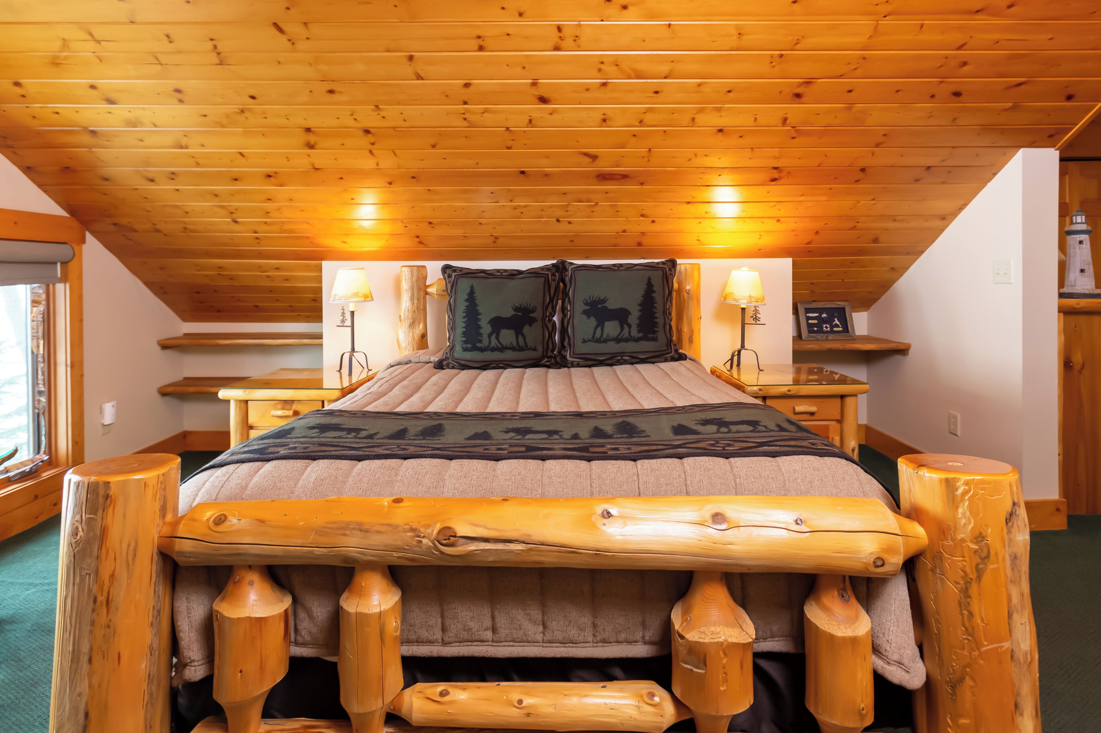 A wooden-framed bed with a headboard, two bedside lamps, and a pillow with a moose design.