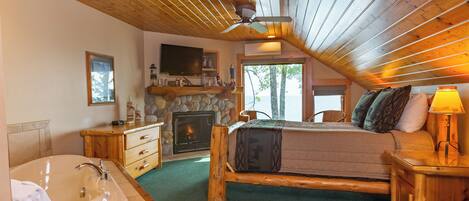 Un dormitorio tipo cabaña de troncos con chimenea de piedra, una cama grande, cabecera de madera, mesita de noche de madera, una lámpara y un televisor montado en la pared.