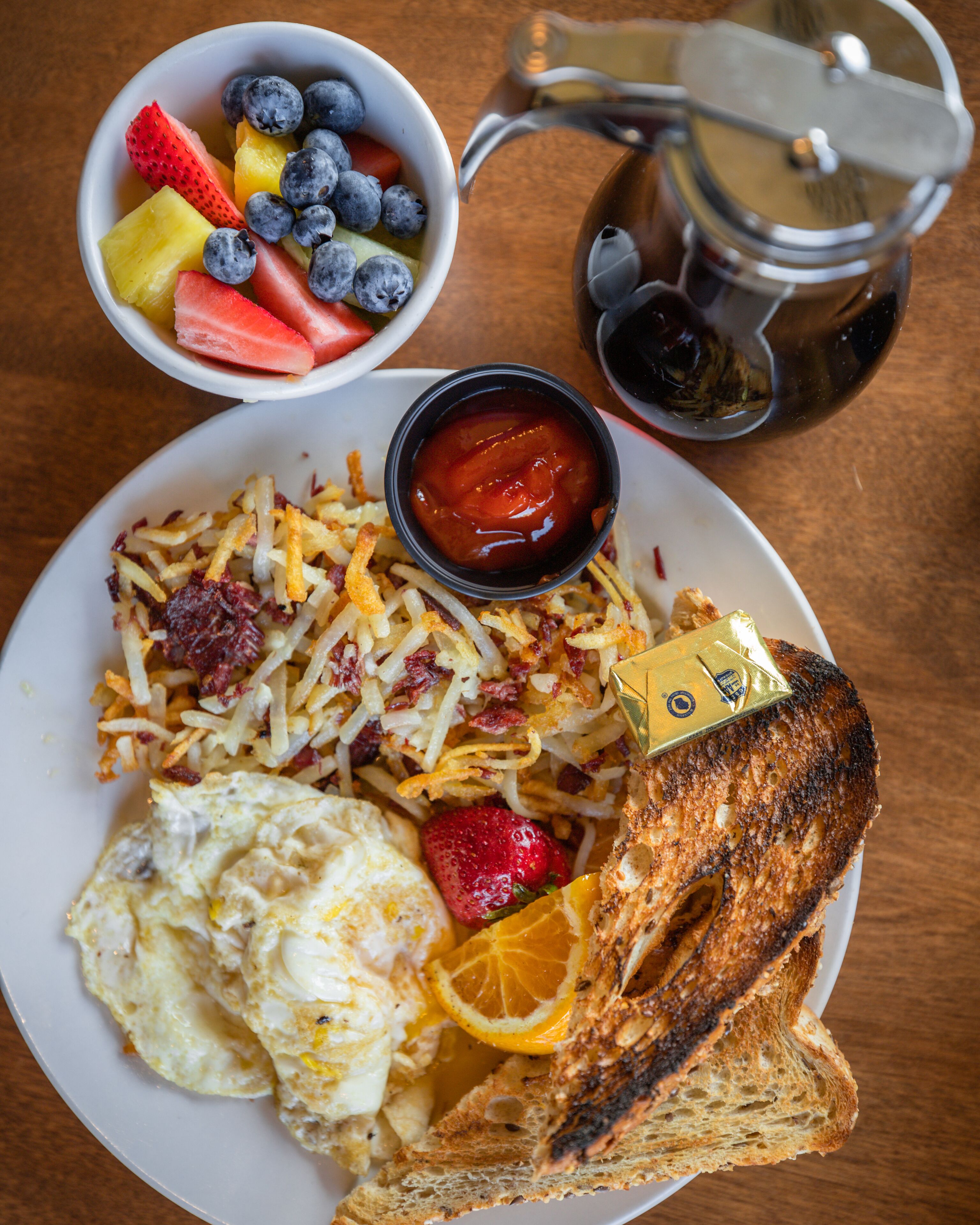 Une assiette de petit-déjeuner comprenant des toasts, des œufs brouillés, des pommes de terre rissolées et un saladier de fruits contenant des fraises, des myrtilles et de la pastèque.