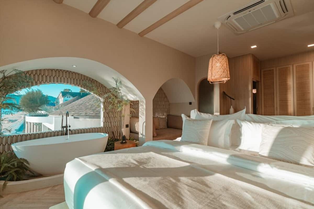 A bedroom with a large bed, a bathtub, and a view of a house through an arched window.