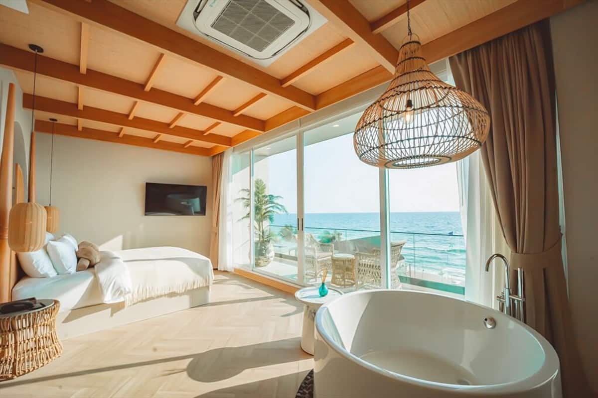 A modern bedroom with a large window, a white bathtub, and a wicker lamp.