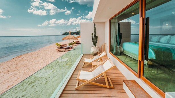 A modern beachside room with a large glass window overlooking the sea, a wooden deck with lounge chairs, and a cactus plant. - Maldives Beach Resort (Tha Mai)
