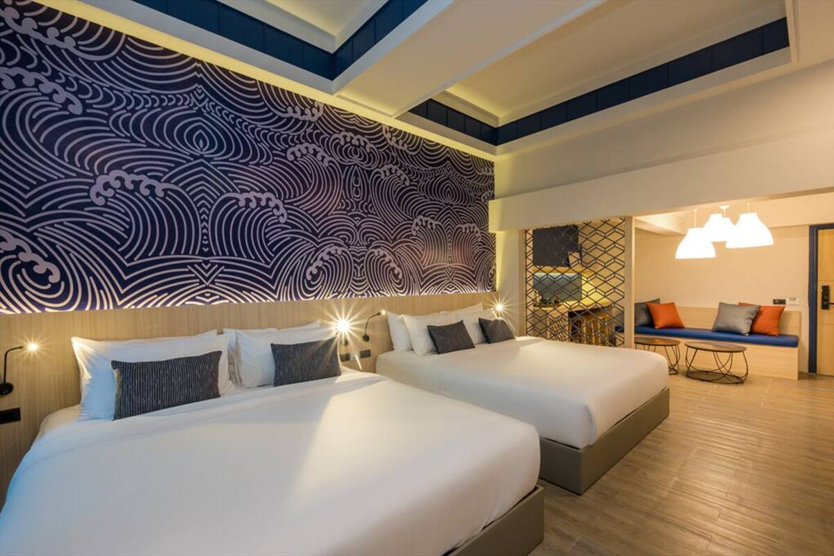 A hotel room with two beds, a decorative wall, a seating area with a sofa and table, and a patterned ceiling.