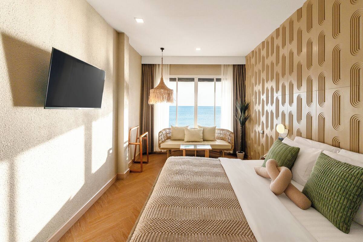 A hotel room with a bed, a TV mounted on the wall, a sofa, a dining table, and a view of the sea.