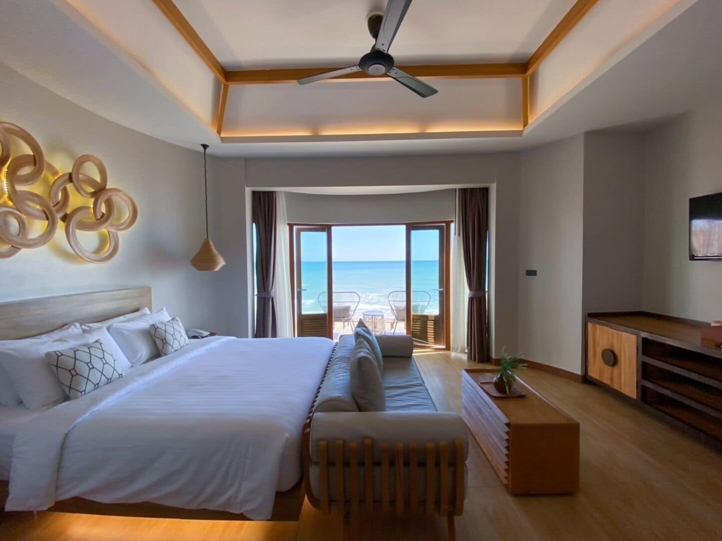 Habitación de hotel con una cama grande, una zona de estar con vista al mar, un ventilador de techo y un televisor.