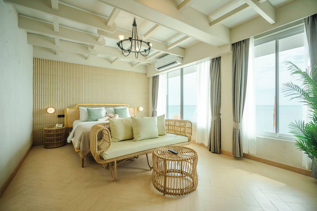 A room with a bed, a wicker sofa, a round wicker coffee table, a chandelier, and large windows with curtains.
