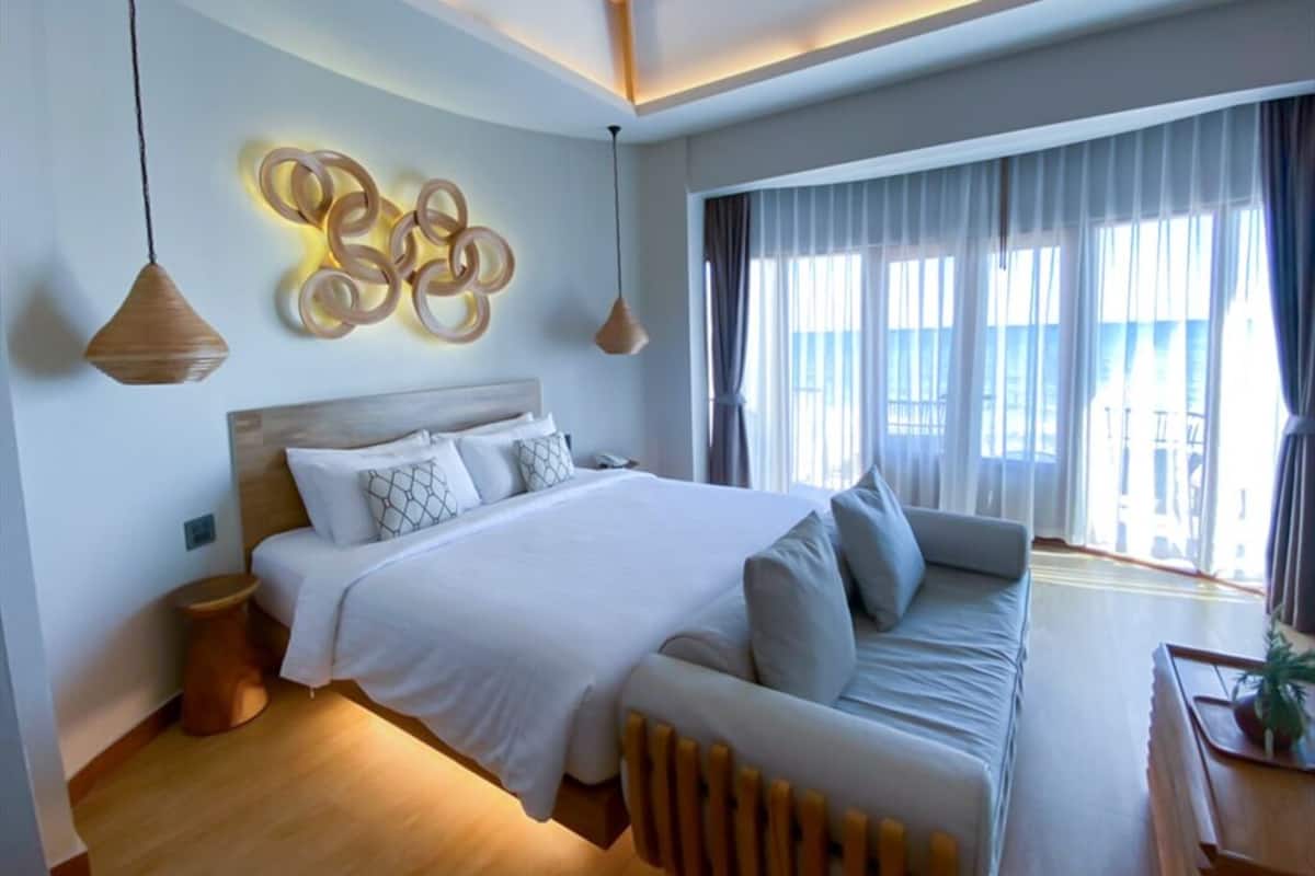 A modern hotel room with a large bed, a sofa, a wooden side table, and a decorative wall piece.