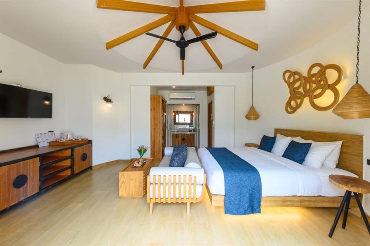 A modern hotel room with a wooden ceiling, a large bed with blue bedding, a wooden side table, a sofa, and a TV mounted on the wall.