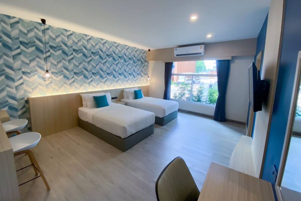 A modern hotel room with two beds, a dining area, and a large window providing a view of the outdoors.