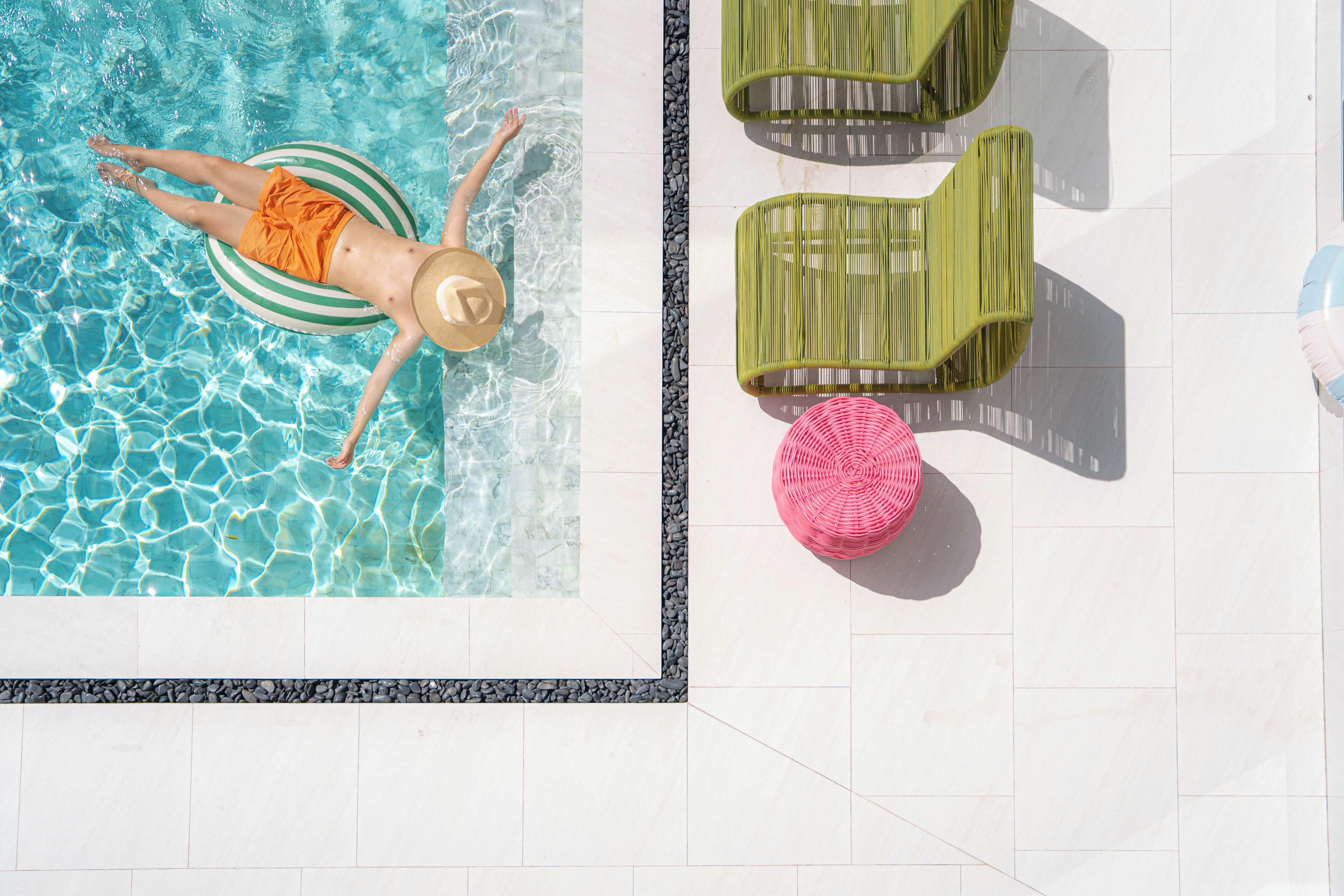 A swimming pool with a person floating on a green and white inflatable ring, surrounded by lounge chairs and a pink woven stool.