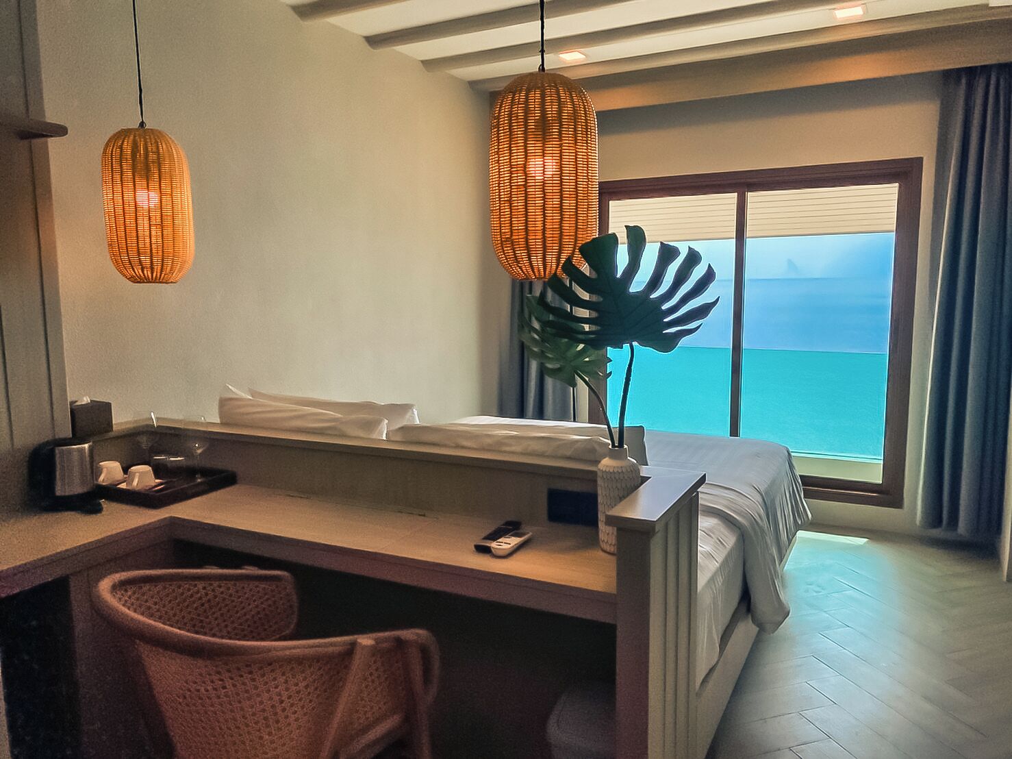 A hotel room with a bed, a desk with a chair, a large window with a view of the sea, and two hanging wicker lamps.