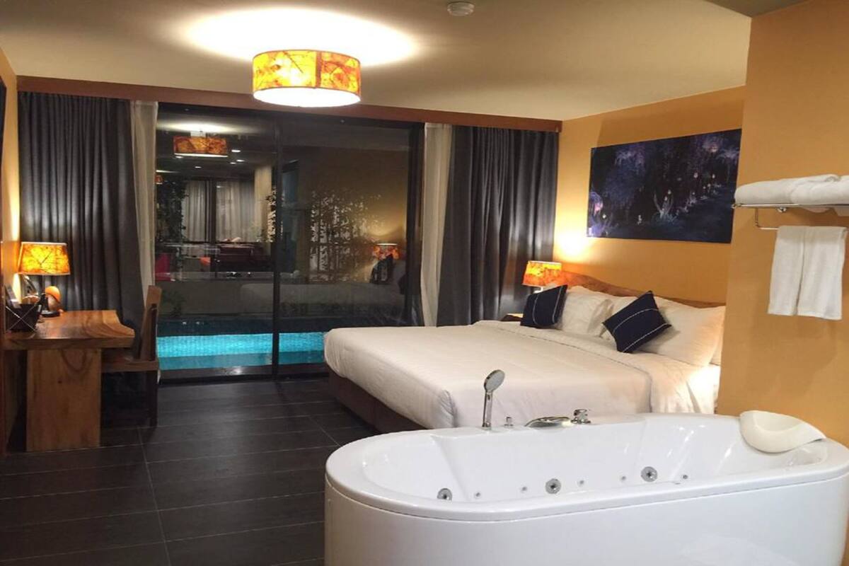 A hotel room with a large bed, a bathtub, a desk with a lamp, and a view of a pool through a glass door.