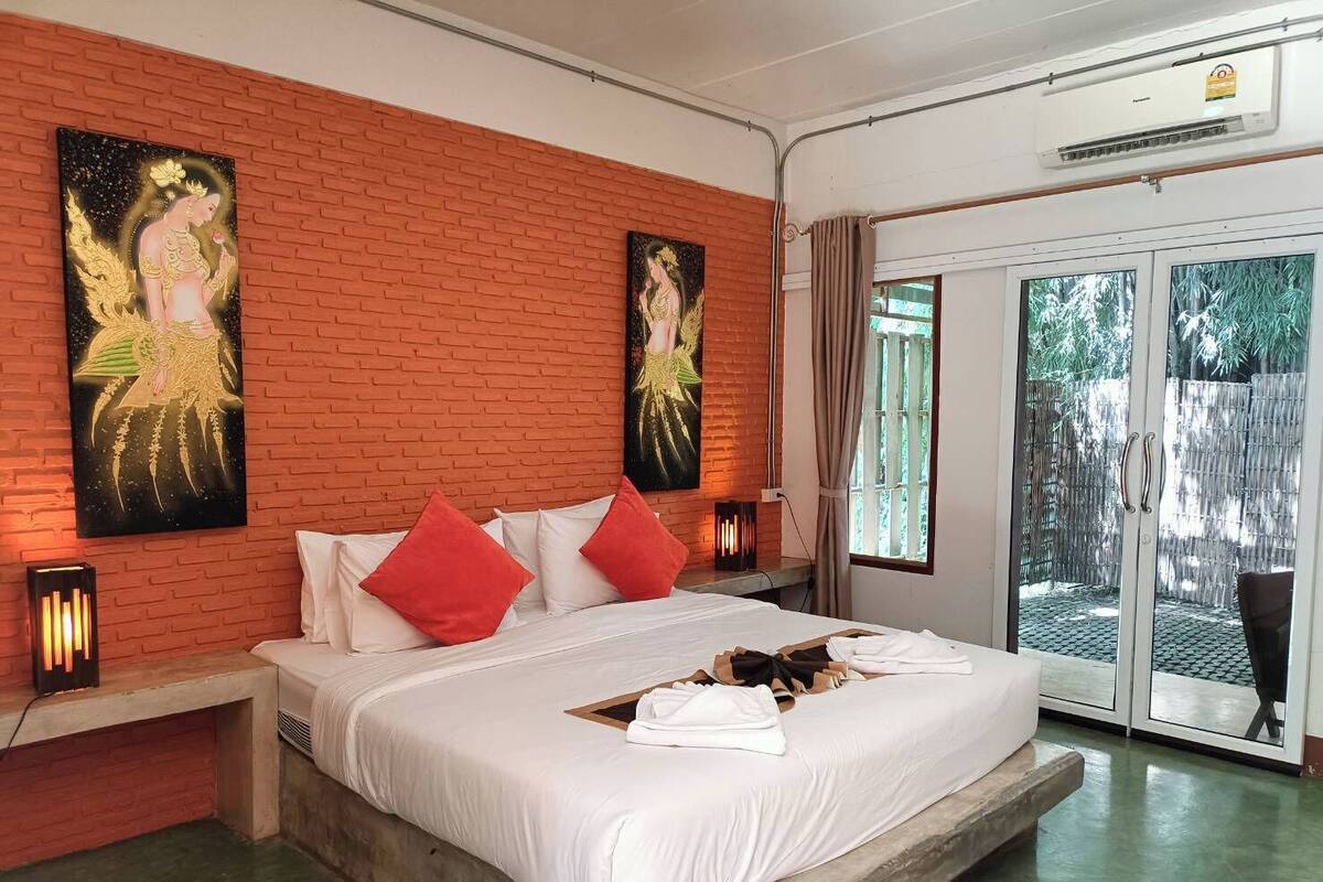 Habitación de hotel con pared de ladrillo, cama con ropa blanca y almohadas rojas, mesita de noche con lámpara y puerta de vidrio corrediza que da a un área exterior.