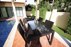 An outdoor patio with a glass-top table and wicker chairs, a tiled pool, and a white wall with greenery. - Bang Saray Pool Villa by Pattaya Sunny Rentals (Sattahip)