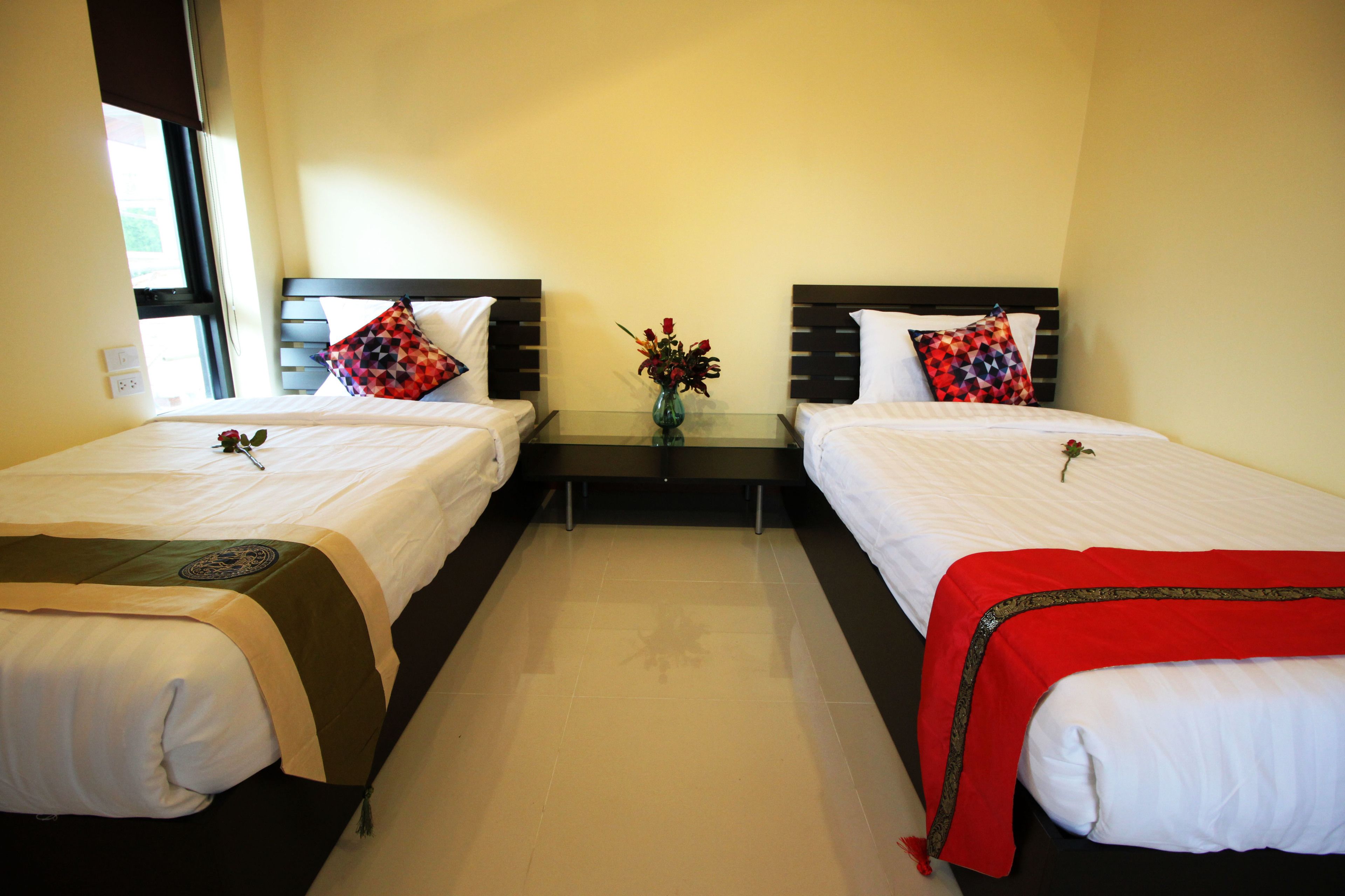 A hotel room with two single beds, a small table with a vase of flowers, and a window providing natural light.