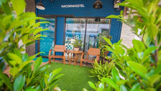 Un Nacornhostel à l’extérieur bleu, doté d’un petit espace extérieur avec des chaises et des tables en bois, et d’une pelouse artificielle verte.