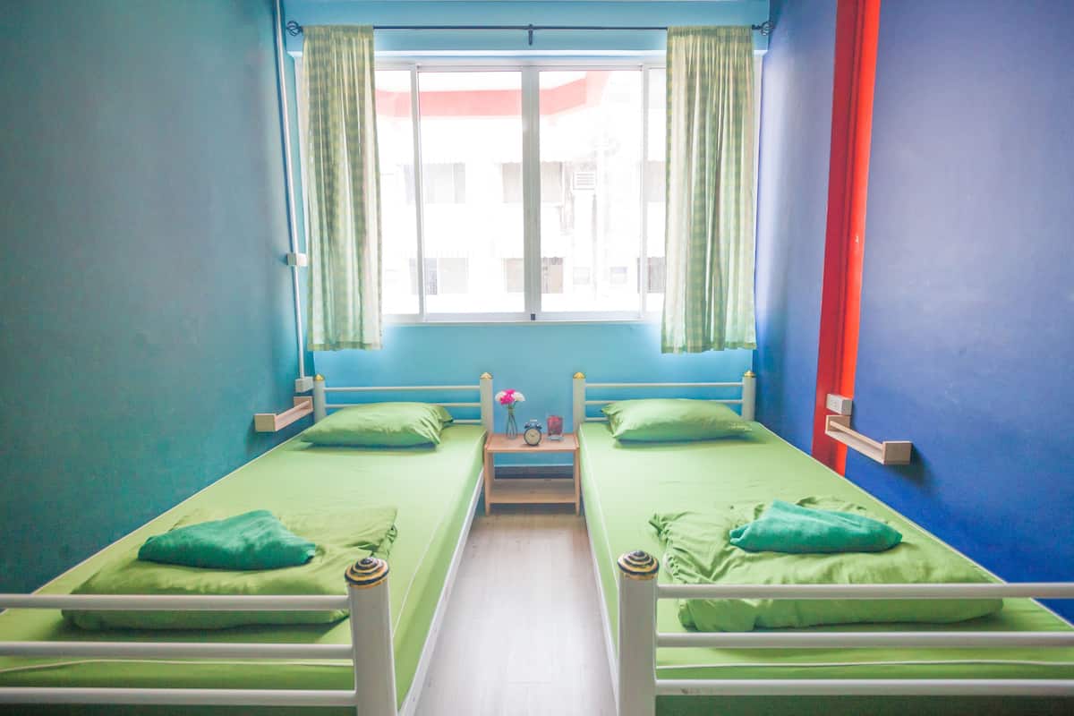 Une chambre avec deux lits simples, une petite table sur laquelle se trouvent une horloge et un vase, et une fenêtre avec des rideaux.