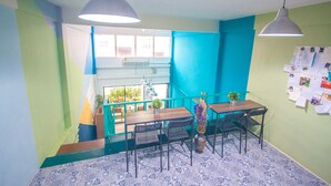 Une pièce avec un escalier, une table entourée de chaises, un luminaire suspendu et un tableau d’affichage recouvert de feuilles de papier.