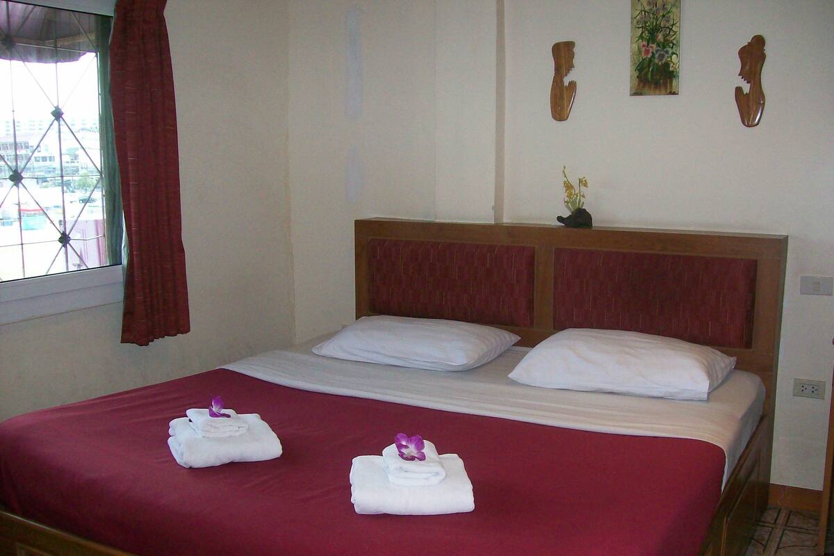 A neatly made bed with a red bedspread and white pillows, two towels folded on the bed, and a window with a view of buildings outside.