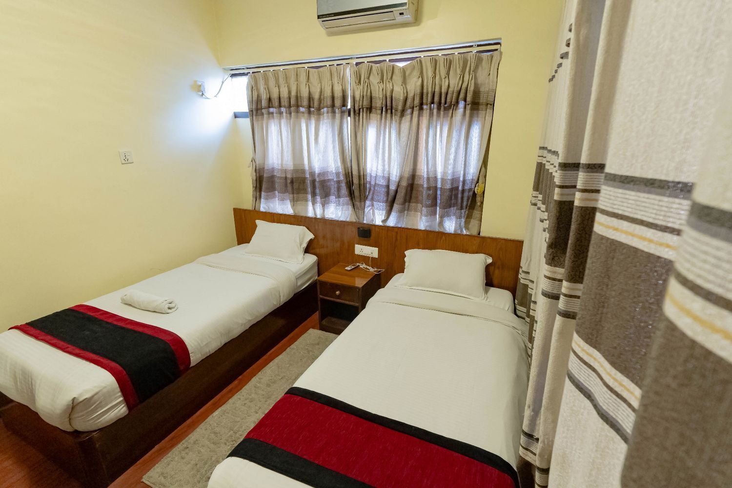 Una habitación con dos camas individuales, cabecera de madera, mesita de noche, teléfono y ventana con cortinas.